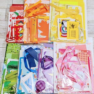 Könnte beinhalten: Eine Sammlung von sechs farblich sortierten Bastelsets, die in einem Raster angeordnet sind. Jedes Set enthält diverses Papier, Bänder, Spielkarten und kleine Verzierungen in Rot, Orange, Gelb, Grün, Blau und Pink, verpackt in Klarsichtfolie.