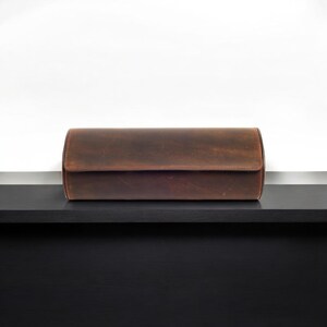 May include: A brown leather watch roll with a cylindrical shape, sitting on a black surface.