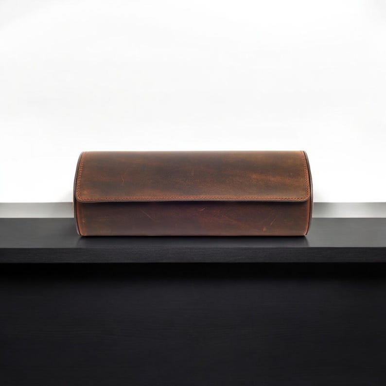 May include: A brown leather watch roll with a cylindrical shape, sitting on a black surface.