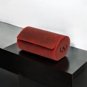 May include: A red leather watch winder with a monogrammed initial "A" and antlers on the side.