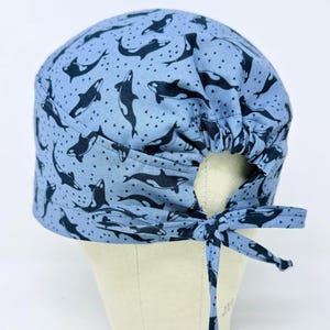 May include: A light blue scrub cap featuring a pattern of black and white orcas. The cap has a gathered back and ties for an adjustable fit. The orca pattern is repeated across the fabric.