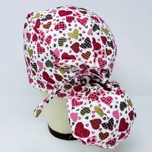 May include: A white scrub hat with a pink, red, and black heart pattern. The hat has a tie at the back that can be tied into a bun.