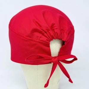 Op de afbeelding: Een felrode scrub cap met een gerimpelde achterkant en een striksluiting. De cap, gemaakt van gladde stof, wordt getoond op een mannequinhoofd. Dit medische accessoire is ontworpen voor zorgprofessionals.