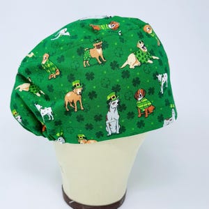 May include: Green surgical scrub cap with a pattern of dogs wearing St. Patrick's Day attire, including green hats and sweaters, and shamrocks. The cap is displayed on a mannequin head.