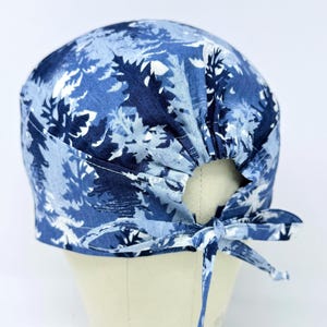 May include: A blue and white patterned scrub cap with a tie closure. The cap features a repeating leaf design in shades of blue and white. The cap is tied in a bow at the back.