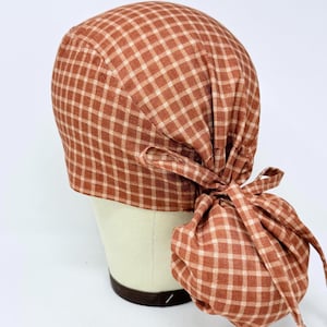 Puede incluir: Una gorra de exfoliación a cuadros de color óxido con un sujetador de cola de caballo a juego con lazo. La gorra está hecha de una tela suave y está diseñada para un ajuste cómodo. El patrón de cuadros es un diseño clásico.