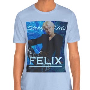 Male model showcasing the unisex, 100% airlume combed & ring-spun cotton t-shirt in baby blue, featuring a graphic design. It shows Felix from Stray Kids in a provocative pose, wearing a structured black suit jacket and oriental-style patterned pants. The background is a multi-toned blue graffiti. From ModeK Vetements.