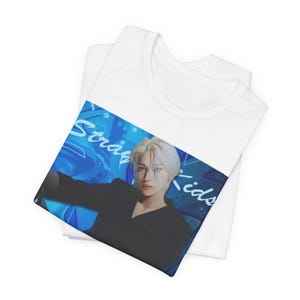 Folded unisex, 100% airlume combed & ring-spun cotton t-shirt in white, featuring a graphic design. It shows Felix from Stray Kids in a provocative pose, wearing a structured black suit jacket and oriental-style patterned pants. The background is a multi-toned blue graffiti. From ModeK Vetements.