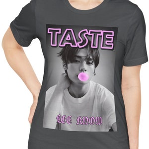 May include: A gray t-shirt with a black and white photo of a young man blowing a pink bubble. The photo is surrounded by a pink neon border with the word "TASTE" in pink neon letters. Below the photo is the text "LEE KNOW" in pink letters.