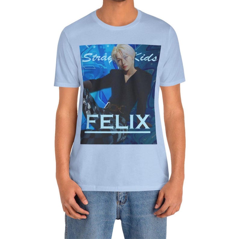 Male model showcasing the unisex, 100% airlume combed & ring-spun cotton t-shirt in baby blue, featuring a graphic design. It shows Felix from Stray Kids in a provocative pose, wearing a structured black suit jacket and oriental-style patterned pants. The background is a multi-toned blue graffiti. From ModeK Vetements.