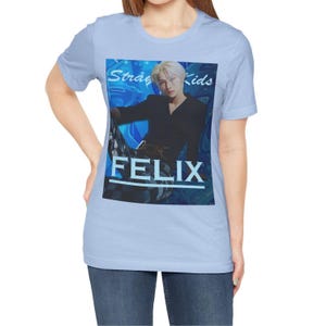 Female model showcasing the unisex, 100% airlume combed & ring-spun cotton t-shirt in baby blue, featuring a graphic design. It shows Felix from Stray Kids in a provocative pose, wearing a structured black suit jacket and oriental-style patterned pants. The background is a multi-toned blue graffiti. From ModeK Vetements.