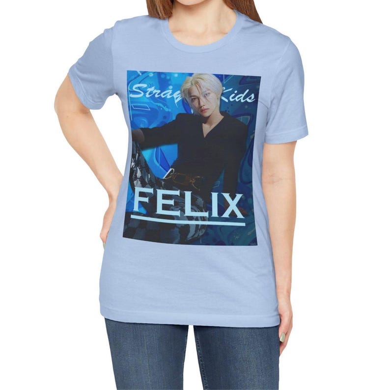 Female model showcasing the unisex, 100% airlume combed & ring-spun cotton t-shirt in baby blue, featuring a graphic design. It shows Felix from Stray Kids in a provocative pose, wearing a structured black suit jacket and oriental-style patterned pants. The background is a multi-toned blue graffiti. From ModeK Vetements.