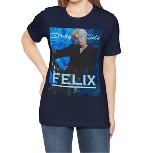 Female model showcasing the unisex, 100% airlume combed & ring-spun cotton t-shirt in navy, featuring a graphic design. It shows Felix from Stray Kids in a provocative pose, wearing a structured black suit jacket and oriental-style patterned pants. The background is a multi-toned blue graffiti. From ModeK Vetements.