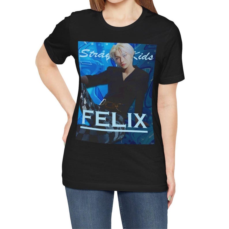 Female model showcasing the unisex, 100% airlume combed & ring-spun cotton t-shirt in black, featuring a graphic design. It shows Felix from Stray Kids in a provocative pose, wearing a structured black suit jacket and oriental-style patterned pants. The background is a multi-toned blue graffiti. From ModeK Vetements.