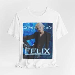 Draped unisex, 100% airlume combed & ring-spun cotton t-shirt in white, featuring a graphic design. It shows Felix from Stray Kids in a provocative pose, wearing a structured black suit jacket and oriental-style patterned pants. The background is a multi-toned blue graffiti. From ModeK Vetements.