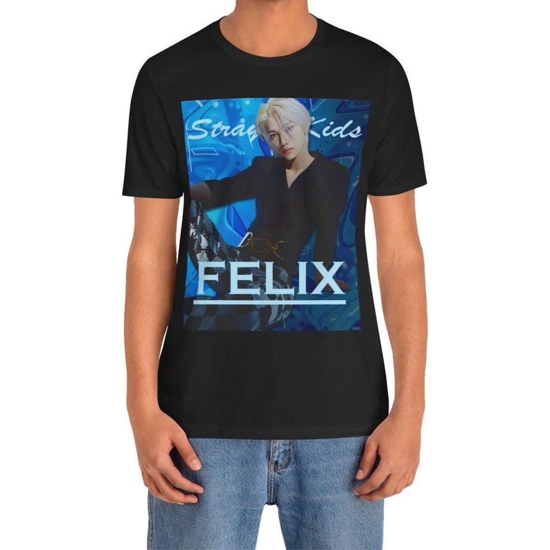 Male model showcasing the unisex, 100% airlume combed & ring-spun cotton t-shirt in black, featuring a graphic design. It shows Felix from Stray Kids in a provocative pose, wearing a structured black suit jacket and oriental-style patterned pants. The background is a multi-toned blue graffiti. From ModeK Vetements.