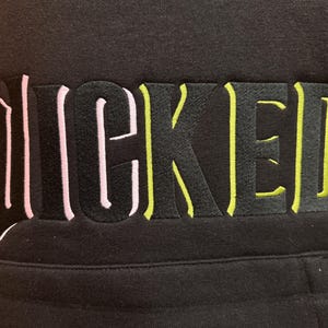 May include: Black hoodie with the word "WICKED" embroidered across the chest. The lettering is black with pink and yellow accents. The hoodie has a front pocket.