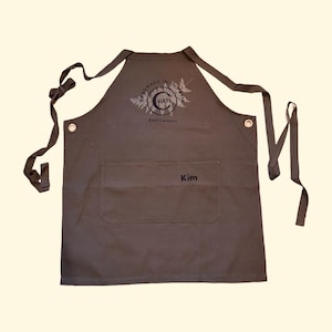 May include: A dark grey apron with a large front pocket and long ties. The apron has the name "Kim" embroidered on the pocket. The top of the apron has a circular logo with the text "MADE IN COLUMBIA" and "KAH Ceramics".