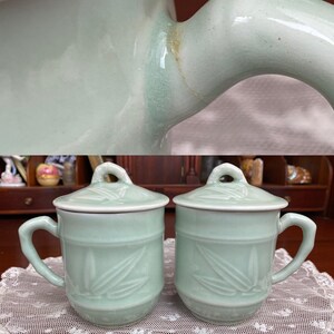 May include: Two matching pale green ceramic mugs with lids. Each mug has a handle and a lid with a small loop. The mugs feature a bamboo leaf design. The mugs are sitting on a white lace tablecloth.