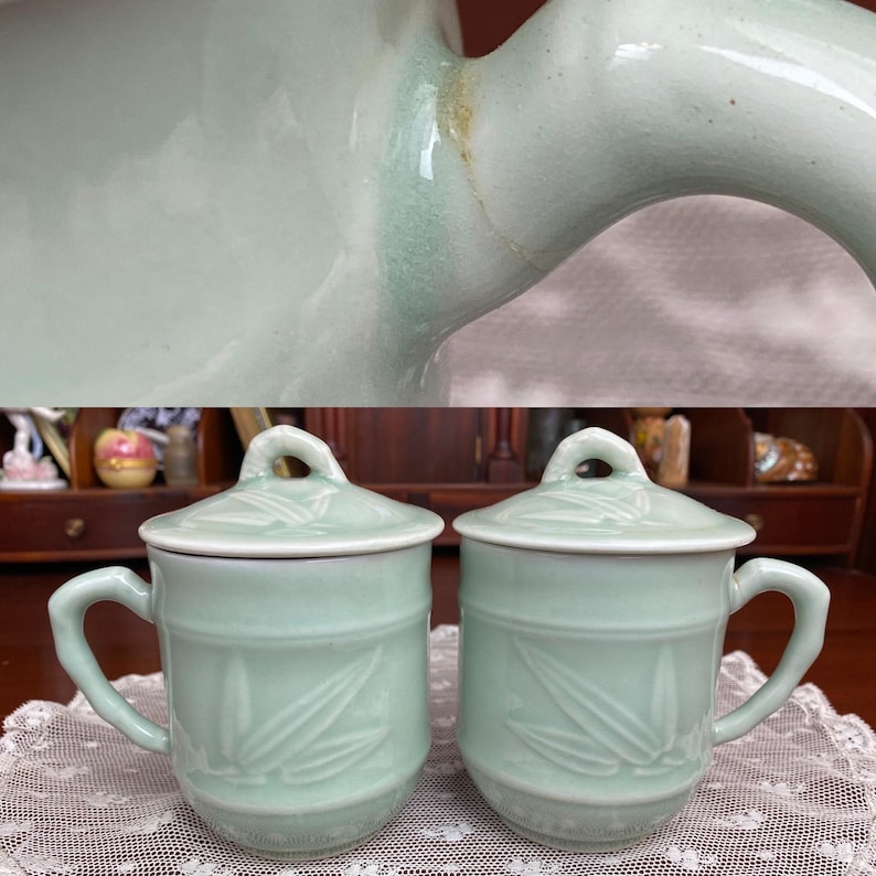 May include: Two matching pale green ceramic mugs with lids. Each mug has a handle and a lid with a small loop. The mugs feature a bamboo leaf design. The mugs are sitting on a white lace tablecloth.