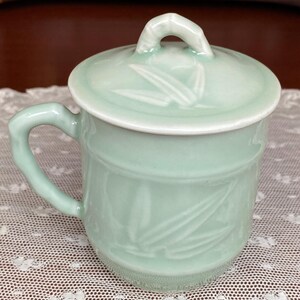 May include: A pale green ceramic mug with a matching lid. The mug has a handle and is decorated with a bamboo leaf design. The lid has a small loop handle. The mug is on a white lace tablecloth.