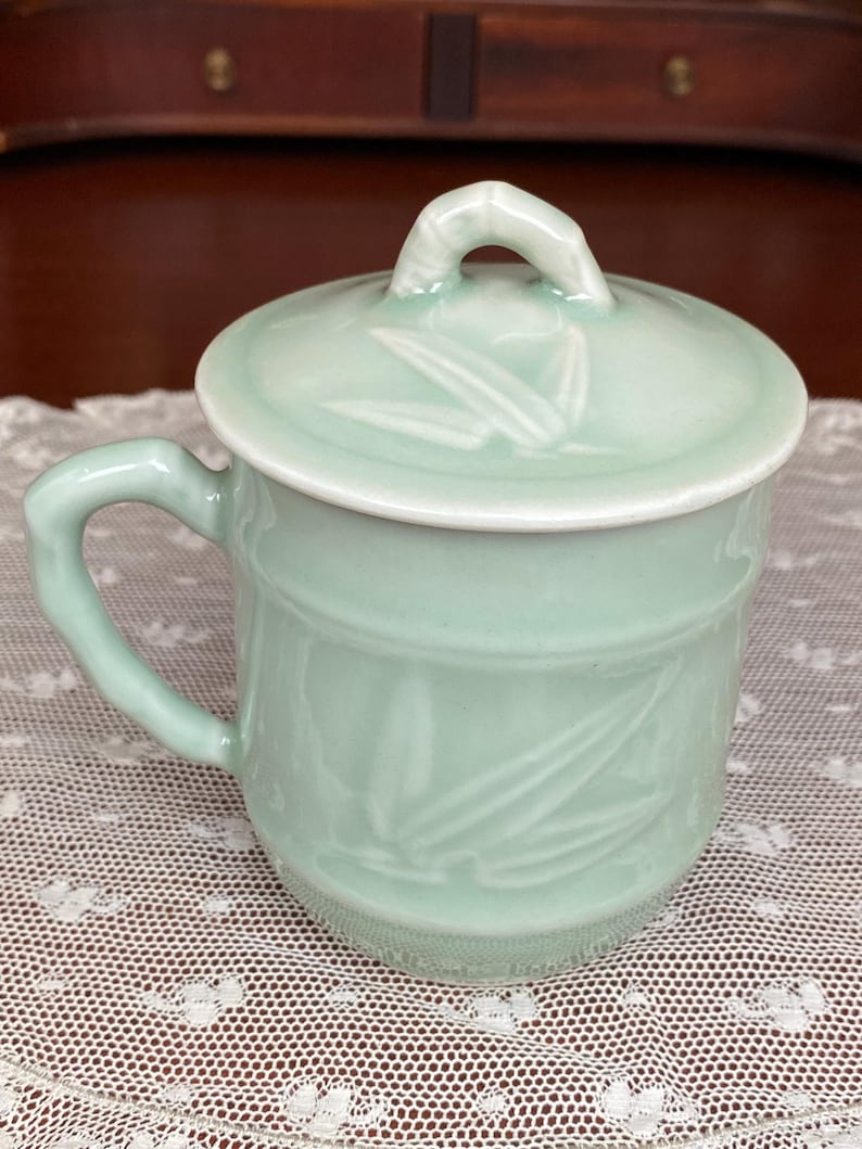 May include: A pale green ceramic mug with a matching lid. The mug has a handle and is decorated with a bamboo leaf design. The lid has a small loop handle. The mug is on a white lace tablecloth.