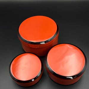 May include: Three round, nesting containers with glossy red lids and black rims. The containers vary in size, with the largest in the back and the smallest in the front. The containers are made of a dark material.