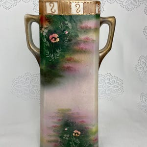 May include: Rectangular vase with a painted floral design. The vase has a light pink and green background with pink flowers and green foliage. The top of the vase has a gold trim with the number "2" on it. The vase has two gold handles.