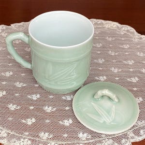 May include: A pale green ceramic mug with a matching lid. The mug has a handle and is decorated with a bamboo leaf design. The lid has a small handle and also features the bamboo leaf design. The mug and lid are sitting on a white lace doily.