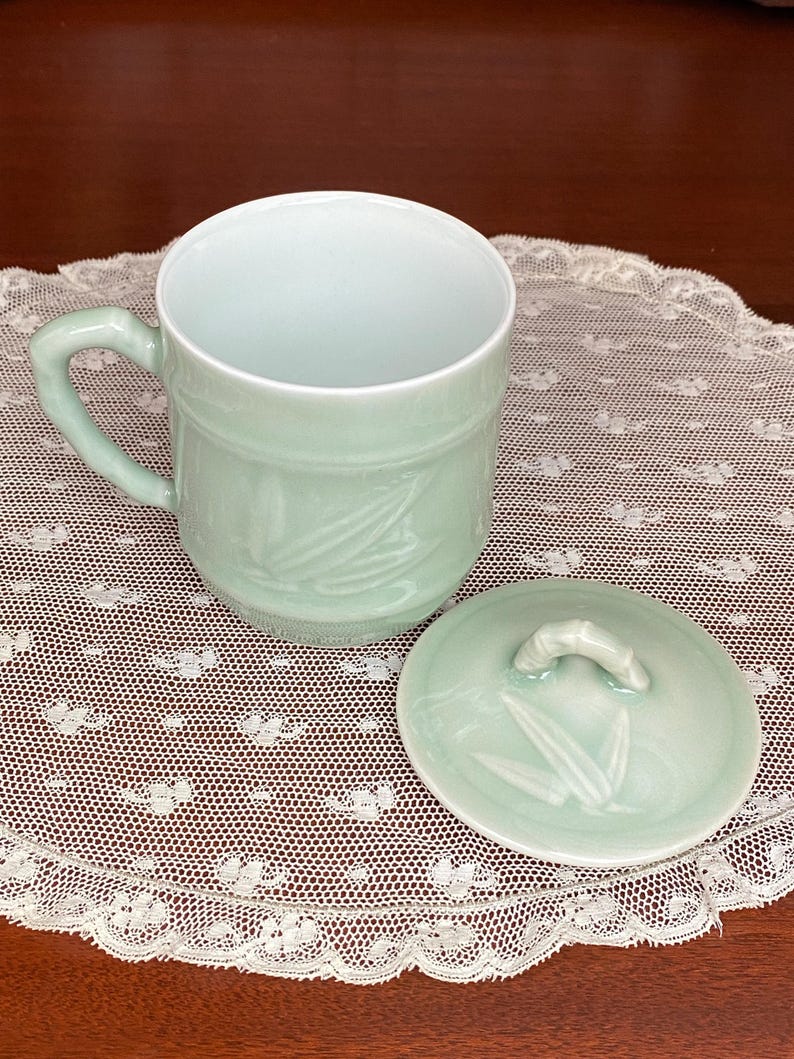 May include: A pale green ceramic mug with a matching lid. The mug has a handle and is decorated with a bamboo leaf design. The lid has a small handle and also features the bamboo leaf design. The mug and lid are sitting on a white lace doily.