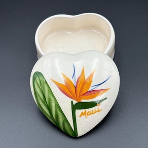 May include: Heart-shaped trinket box with a lid featuring a hand-painted bird of paradise flower in orange, blue, and green. The word "Maui" is written in orange script. The box is off-white.