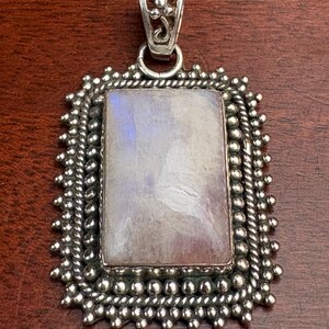May include: A silver pendant with a rectangular, iridescent moonstone center. The stone is set within a detailed, beaded frame. The bail features an ornate, openwork design. The pendant is displayed on a dark wood surface.