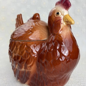 May include: A ceramic hen-shaped container with a lid. The hen is primarily brown with a glossy finish, featuring a red comb and yellow beak. The container is designed to resemble a sitting hen, with detailed feather textures.