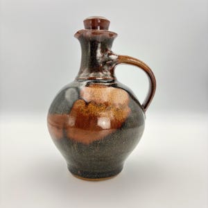 May include: A ceramic cruet with a rounded body, a narrow neck, and a single handle. The piece features a glossy glaze with mottled patterns of deep black, dark brown, and warm amber tones. A matching ceramic stopper sits in the spout opening.