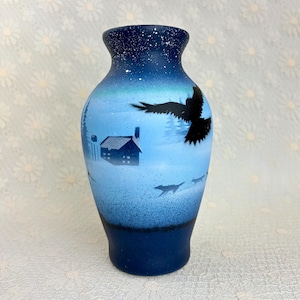 May include: A blue vase with a winter scene. The vase features a dark blue base that fades to a lighter blue with a snowy night sky design. A black silhouette of a bird in flight and a house are also featured.