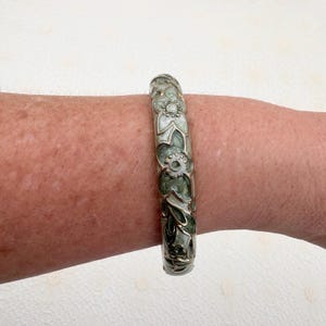 May include: A rigid bangle bracelet featuring a repeating floral pattern in shades of sage green and cream. The design is accented with metallic silver-toned outlines that define the petals and leaves, creating a textured, raised appearance.