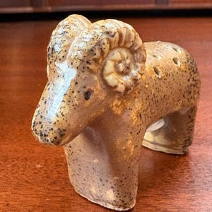 May include: A ceramic ram figurine with a speckled tan and brown glaze. The ram features prominent curled horns, a smooth snout, and a series of small circular holes along its back. The piece has a glossy finish and stands on four legs.
