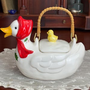 May include: A white ceramic duck-shaped cookie jar with a red scarf and a yellow beak. The lid is topped with a small yellow chick figurine, and it has a woven handle. The jar is decorated with green holly leaves.