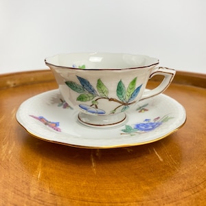 May include: A white porcelain teacup and saucer with a floral design. The cup has a scalloped rim and a gold trim. The saucer has a delicate floral pattern and a gold rim.