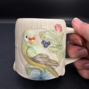 May include: Cream-colored ceramic mug with a raised relief design of a parrot, flowers, and foliage. The parrot is primarily green with a red beak and is perched on a blue and yellow base. The mug has a handle and vertical ribbed detailing.