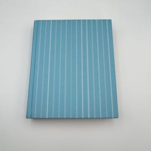 Puede incluir: Un cuaderno rectangular azul claro con l&iacute;neas de puntos blancas verticales. El cuaderno est&aacute; cerrado y tiene una cubierta texturizada. El lomo tambi&eacute;n es azul claro.