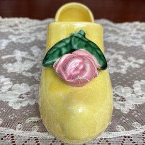 May include: A yellow ceramic shoe-shaped vase with a pink rose and green leaves. The vase has a small opening at the top and is decorated with a floral design. The shoe is a decorative item.