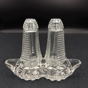 May include: Clear glass salt and pepper shakers with a matching tray. The shakers have a rectangular shape with vertical lines and a domed top. The tray has a decorative, scalloped edge.