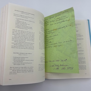 Puede incluir: P&aacute;ginas de libro de cocina abiertas con recetas de espinacas. Una nota adhesiva verde con una receta manuscrita est&aacute; insertada. El libro est&aacute; sobre una superficie blanca.