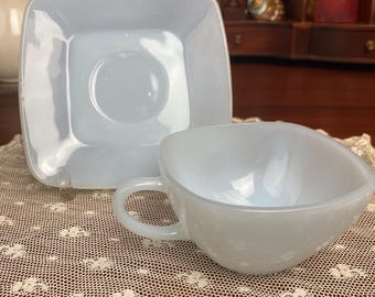 May include: A white milk glass teacup and saucer set. The cup has a rounded shape and a handle. The saucer is square with a slightly raised edge.