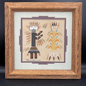 May include: Framed sand painting featuring two stylized figures in earthy tones. The artwork, set against a light background, is framed in a natural wood. The figures have geometric designs and are likely of Native American origin.