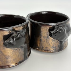 May include: Two ceramic mugs with a bronze and black finish. Each mug features a sculpted pig head as a handle. The mugs have a rustic, handmade appearance, with a metallic sheen. The interior of the mugs is dark brown.