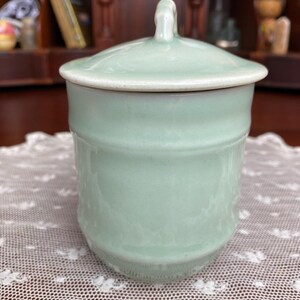 May include: A pale green ceramic jar with a matching lid. The jar has a cylindrical shape with a slightly textured surface and a white rim. The lid features a small handle. The jar is on a white lace tablecloth.