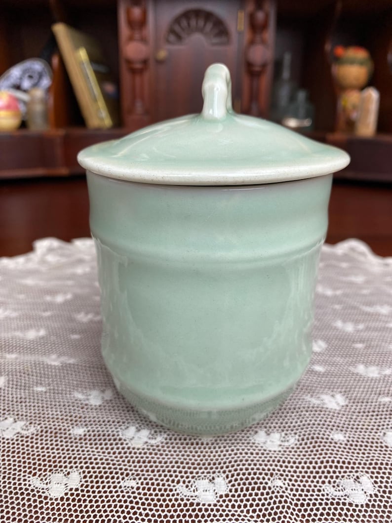 May include: A pale green ceramic jar with a matching lid. The jar has a cylindrical shape with a slightly textured surface and a white rim. The lid features a small handle. The jar is on a white lace tablecloth.