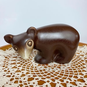 May include: A brown ceramic bear figurine with white accents around the eyes and nose. The bear is sitting on a white crocheted doily.